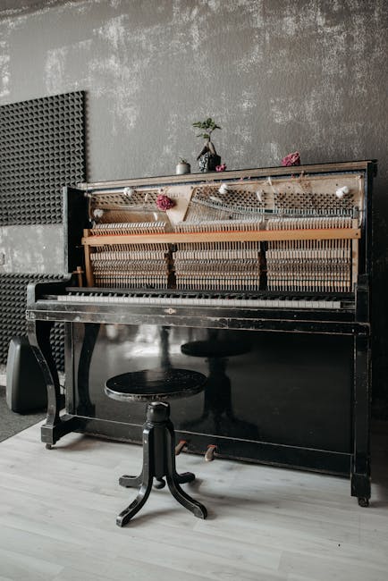 A vintage black grand piano with an open lid revealing the interior, positioned inside a room with weathered brick and ornate, aged wall decorations. In front of the piano is a small, round, padded st