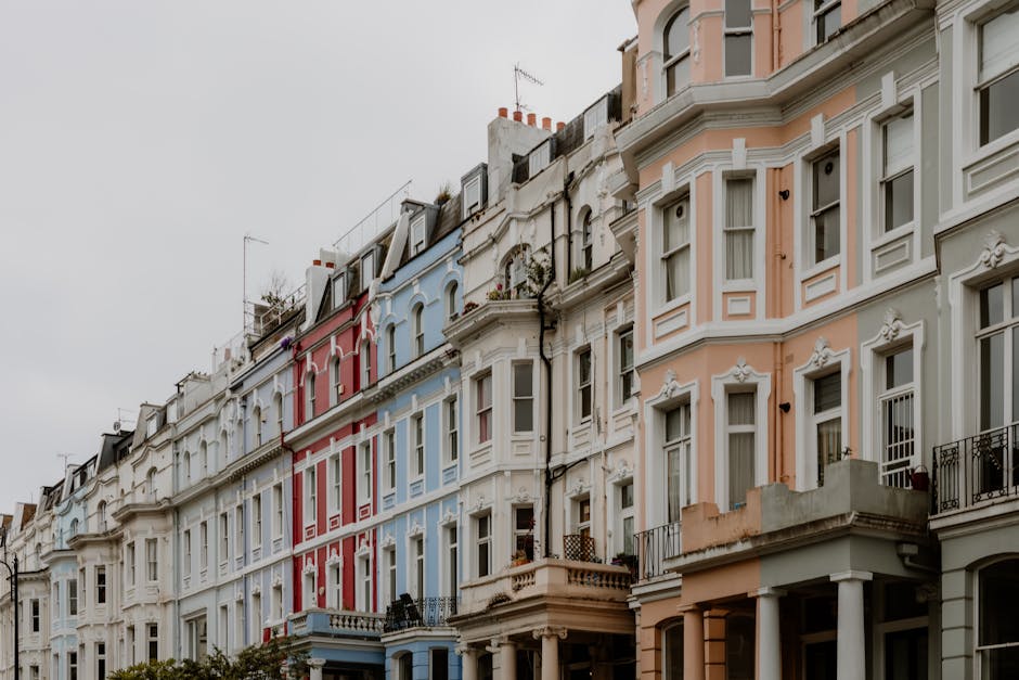 A row of multi-storey residential buildings painted in light blue and beige tones, with decorative architectural details such as bay windows, balconies, and columns. The buildings are situated along a