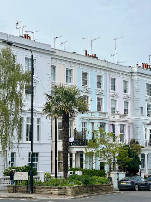 A row of white Victorian terraced houses with multiple stories, large sash windows, and decorative architectural detailing, situated on a street with a pavement. In front of the buildings, there are various trees, including a tall palm tree and a weeping willow, as well as small shrubs and garden beds enclosed by low brick walls. A black lamppost is visible near the pavement. A dark grey car is parked along the street, close to the entrance of one of the houses. The sky above the buildings is overcast with a few visible antennas on the rooftops. This residential area is typical of Notting Hill, with well-maintained facades and greenery. The scene is captured during daylight, illustrating the exterior environment where house removals and furniture transport may occur as part of a home relocation process supported by [COMPANY_NAME], as described in the 'Ladbroke Grove moves: navigating narrow Victorian stairs' page on moversnottinghill.co.uk.