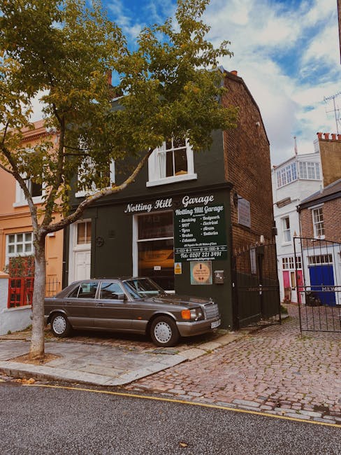 The image shows a narrow cobbled street in Notting Hill featuring a two-storey building with dark green exterior walls and a white signboard displaying the name 'Notting Hill Garage' alongside various moving and repair services. A classic grey car is parked directly in front of the garage, partially covering the signage. In the foreground, a tree with green leaves is visible, casting slight shadows on the pavement. To the right, there is a black metal gate leading into the property, and to the left, a red-painted fence borders the neighboring building. The street is lined with other residential and commercial buildings, some with brick facades and bay windows, under a partly cloudy sky. This scene captures a typical urban setting where houses and garages are aligned close together, reflecting the compact nature of Notting Hill streets suitable for house removals and furniture transport services provided by Movers Notting Hill.