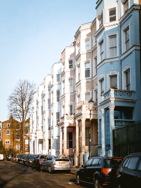 A row of white and light blue Victorian-style terraced houses with ornate architectural details and bay windows lining a narrow street in Notting Hill, with several parked cars along the curb. In the foreground, with a focus on the moving process, there are moving vans parked on the pavement near the property entrances, and a person is visible loading or unloading boxes and furniture covered in protective blankets using trolleys and straps. The vehicles are positioned close to the buildings, indicating the loading or staging area for a home relocation. The scene is illuminated by natural daylight, with a clear sky and some trees visible in the background, reflecting an urban setting typical of house removals and furniture transport operations carried out by Movers Notting Hill, supporting seamless packing and moving logistics involved in the service category.