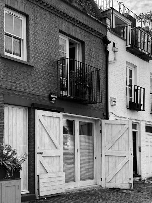 The image shows a narrow cobbled street in Notting Hill featuring a two-storey building with dark green exterior walls and a white signboard displaying the name 'Notting Hill Garage' alongside various moving and repair services. A classic grey car is parked directly in front of the garage, partially covering the signage. In the foreground, a tree with green leaves is visible, casting slight shadows on the pavement. To the right, there is a black metal gate leading into the property, and to the left, a red-painted fence borders the neighboring building. The street is lined with other residential and commercial buildings, some with brick facades and bay windows, under a partly cloudy sky. This scene captures a typical urban setting where houses and garages are aligned close together, reflecting the compact nature of Notting Hill streets suitable for house removals and furniture transport services provided by Movers Notting Hill.
