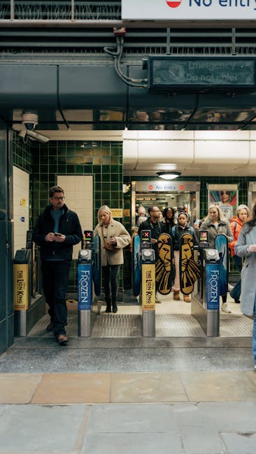 Inside Notting Hill Gate station, a group of commuters wait in front of the ticket barriers and entrance area, which has green tiled walls and a digital display board. The entrance features electronic ticket gate machines with blue and yellow branding, situated on a metal platform with safety railings. Overhead, there is a large ventilation duct, pipes, and electrical wiring running along the ceiling. The station's lighting is bright, illuminating the scene and the varied clothing of the waiting passengers, some of whom are checking their phones or holding bags. The station's environment suggests a typical busy London underground setting, with visible signage indicating station guidelines. As part of the overall context of house removals and moving logistics, the scene highlights the importance of efficient entry and exit points for commuters and relocating individuals transferring between home and transport services, relevant to [PAGE_TITLE], with [COMPANY_NAME] providing professional assistance in aspects of moving and furniture transport.