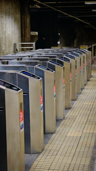 A row of stainless steel turnstiles with electronic card readers at the entrance of Notting Hill Gate station, visible in an underground area with a dark ceiling and concrete walls. The turnstiles are positioned on a tiled floor with a yellow tactile paving strip guiding passengers. In the background, there are a few blurred figures and station lighting, illustrating a typical entry point for commuters. This image reflects the location's infrastructure for managing passenger access, relevant to home relocation or moving services associated with station moves, such as those offered by moversnottinghill.co.uk, especially in the context of efficient transit during furniture transport and packing during house moves.