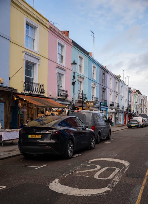 The image displays the exterior of a small, dark green building with a sign reading 'Notting Hill Garage' on its facade, situated on a cobbled street in Notting Hill. In front of the building, a vintage grey car is parked perpendicular to the pavement, partially on the cobblestone area, close to a mature tree with green foliage. The building has large front windows with white frames and a door to the right, with an additional sign listing services such as removals, garage services, and storage. The surrounding environment includes neighboring residential buildings with red brick and white facades, some with bay windows and balconies. The sky is partly cloudy with patches of blue, and the scene suggests a typical street scene during daytime, relevant to home relocation and furniture transport, with potential involvement of loading and unloading activities by professional moving services like Movers Notting Hill.