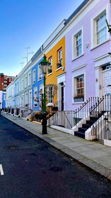 The image shows a row of colorful terraced houses on a residential street in Notting Hill, with facades painted in pastel shades including blue, yellow, and lavender. Each house has white-framed windows and a small front door with steps leading up from the pavement, some featuring decorative railings and small garden areas. A traditional-style black street lamp is positioned in the foreground near the curb, casting light on the sidewalk. The street appears quiet with no vehicles or pedestrians visible, and the sky is clear with bright daylight illuminating the scene. This setting reflects the characteristic neighborhood in Notting Hill, often associated with house removals and relocation logistics, as a professional moving service might be preparing to load furniture, boxes, and packing materials into a vehicle. The image emphasizes the charming residential environment typical for home relocations in the W11 postcode area.