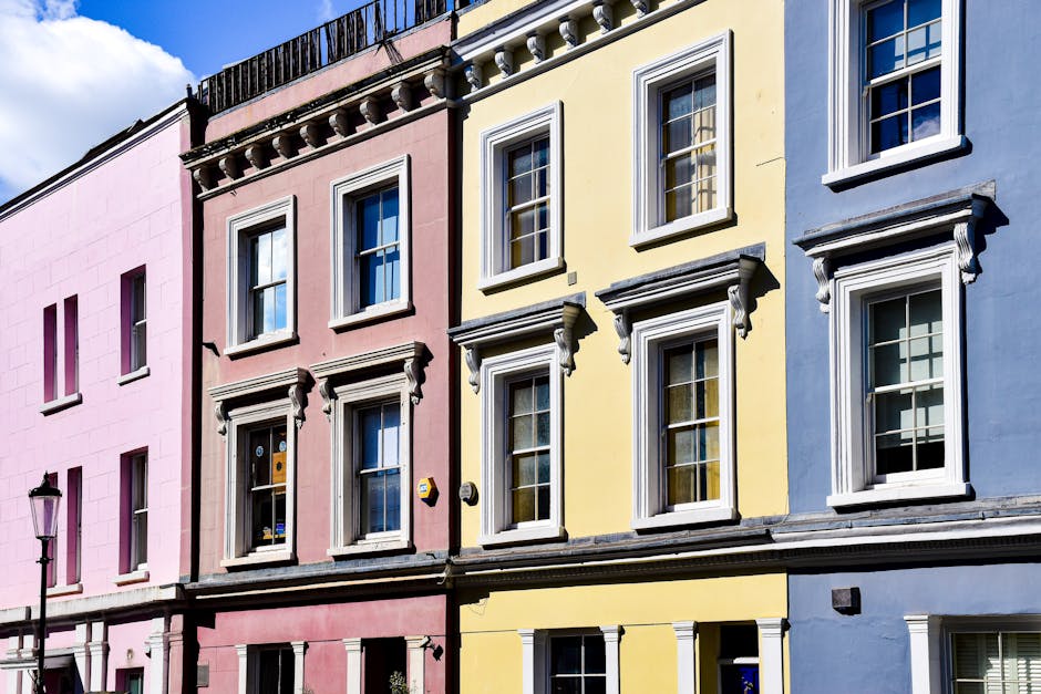 Photograph of a row of three colourful residential buildings on a city street, each featuring classic Victorian architectural details such as decorative window frames and cornices. The left building is painted pink with four visible windows, the middle building is painted yellow with five windows, and the right building is painted blue with four windows, all with white frames and sills. The buildings are situated close together, with a black streetlamp visible on the left side of the image. The scene is taken during daylight with clear blue sky and some clouds overhead. This setting illustrates typical Notting Hill townhouses, which may be involved in home relocation or furniture transport processes carried out by [COMPANY_NAME], highlighting the urban environment associated with house removals and packing and moving activities. The image emphasizes the exterior façades that movers might access during a professional house removal in the W11 postcode area, assisting in visualising the initial phases of a relocation project.