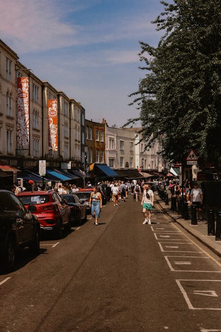 A busy street scene in Notting Hill with parked cars along the left side and a line of multi-story buildings featuring shops and cafes with awnings on the ground level. Pedestrians walk along the pavement, with some carrying boxes or bags, indicating a home relocation process. Towards the right side, a black van is partially visible, likely used for moving furniture and boxes, with the rear doors open. The pavement has designated parking and loading zones, and a large leafy tree extends over part of the sidewalk, providing shade. The image is taken during daylight with a clear sky, illustrating the typical environment where professional removals services by Movers Notting Hill operate, including loading and logistical coordination for house moves in the local area.