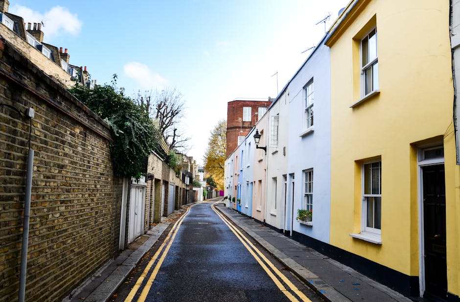 A narrow residential street in Notting Hill with a slightly wet asphalt road marked by double yellow lines on both sides. On the left, a tall, weathered brick wall runs alongside private garages and garden entrances, some partially covered with green foliage. On the right, a row of terraced houses painted in pastel shades of yellow, blue, and white, with white-framed windows and flower boxes. Overhead, a black iron street lamp is attached to a white-painted building, and the sky is partly cloudy with patches of blue. The street appears quiet, suitable for home relocation or furniture transport preparations. This scene captures the typical environment where professional removals like those offered by Movers Notting Hill operate, often navigating these urban spaces during packing and loading processes for house removals.