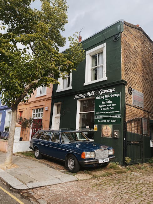 On a quiet street in Notting Hill, a two-storey building housing Notting Hill Garage is visible. The building has a dark green facade with white window frames and a prominent sign displaying the garage's name and contact details. A vintage dark blue estate car with a roof rack is parked directly in front of the garage, partially on a paved sidewalk and a cobblestone street. To the left of the garage, there is a leafed tree with green foliage extending into the frame, providing partial shade. The building’s entrance is on the left side, with a small white door and a narrow window beside it. The area appears well-maintained, and the surroundings include neighboring residential structures with pastel-colored facades. The scene illustrates typical urban surroundings where professional house removals or furniture transport services, such as those provided by Movers Notting Hill, can be carried out efficiently, especially during home relocations or packing and moving operations involving loading and unloading furniture and packing materials.