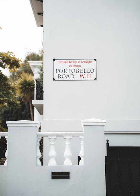 The exterior of a white residential building on Portobello Road in the W11 postcode area is visible, showing a section of a white wall with a decorative balustrade made of white-painted concrete or stone, positioned at the front of the property. A black mailbox is attached to the wall beneath the balustrade, and a small part of a black metal gate is seen on the right side of the image. Above the wall, a white sign with red and black lettering indicates the location as in the Royal Borough of Kensington and Chelsea, specifically on Portobello Road, W11. The background features blurred greenery and trees, suggesting the property is in a leafy area. The scene is lit with natural daylight, with soft shadows cast on the building surface. This image relates to house removals and furniture transport, illustrating an urban property possibly involved in packing, loading, or moving logistics managed by moversnottinghill.co.uk, who specialise in home relocation services in Notting Hill.