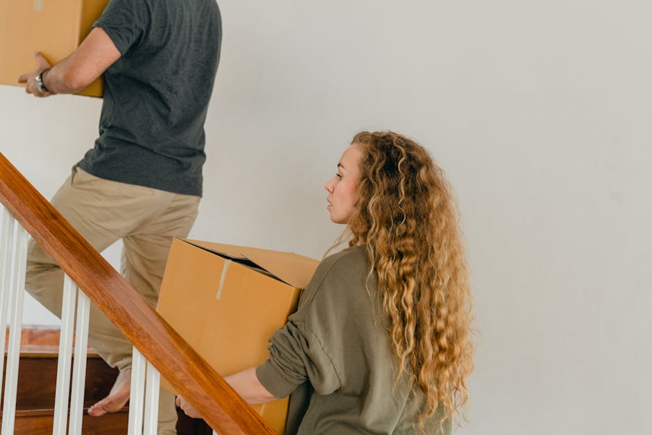 A woman with long, curly red hair and light skin carrying a medium-sized cardboard box while walking down a staircase inside a residential property. She is dressed in an olive green top with sleeves rolled up. The staircase has wooden steps and white-painted balustrades. In the background, a man with light skin in a dark grey t-shirt and beige trousers is ascending the stairs, holding a similar cardboard box. Both are engaged in a home relocation or packing process, facilitated by Movers Notting Hill, as part of a furniture transport or removals service. The environment is well-lit with natural light, and the scene depicts a typical interior space during a house move, emphasizing careful handling of packed boxes and stairs' spatial context during the loading or unloading process.