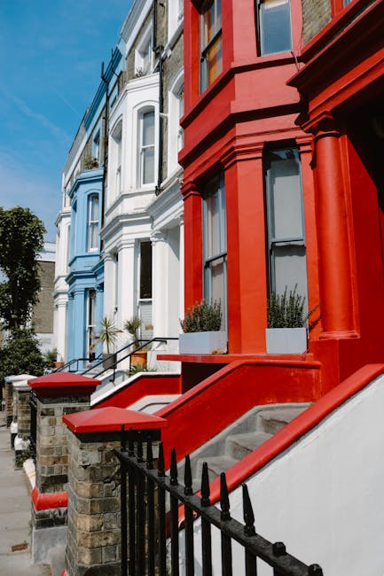 The exterior of a white residential building on Portobello Road in the W11 postcode area is visible, showing a section of a white wall with a decorative balustrade made of white-painted concrete or stone, positioned at the front of the property. A black mailbox is attached to the wall beneath the balustrade, and a small part of a black metal gate is seen on the right side of the image. Above the wall, a white sign with red and black lettering indicates the location as in the Royal Borough of Kensington and Chelsea, specifically on Portobello Road, W11. The background features blurred greenery and trees, suggesting the property is in a leafy area. The scene is lit with natural daylight, with soft shadows cast on the building surface. This image relates to house removals and furniture transport, illustrating an urban property possibly involved in packing, loading, or moving logistics managed by moversnottinghill.co.uk, who specialise in home relocation services in Notting Hill.